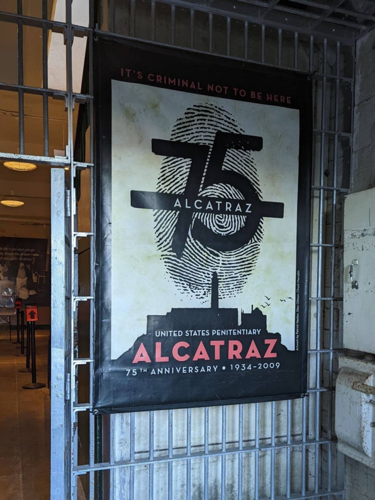 Une affiche noire, blanche et rouge sur la grille d’entrée de la prison d’Alcatraz. Elle promeut le 75ème anniversaire de la prison d’Alcatraz, de 1934 à 1919.