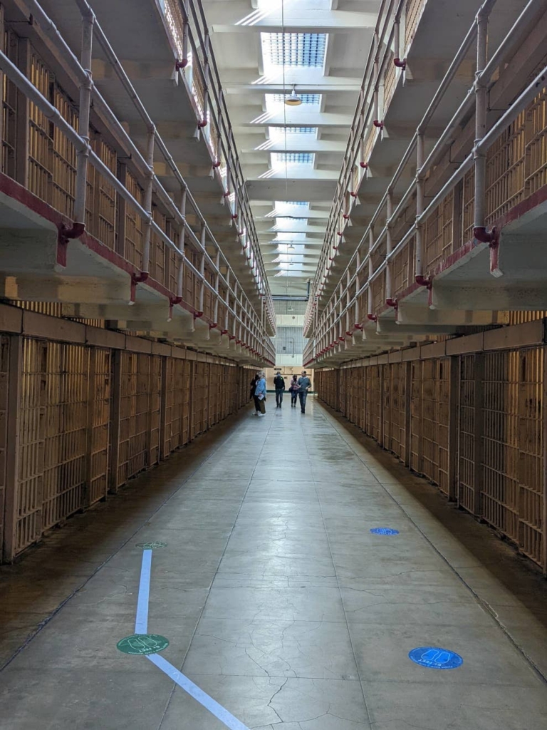 Allée principale de la prison d’Alcatraz. Des cellules sont alignées à droite et à gauche, sur 3 étages. L’allée est immense et très éclairée.