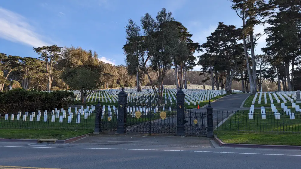 Entrée du cimetière, située de l’autre côté de la route. On peut y observer des tombes blanches et beaucoup d’arbres.