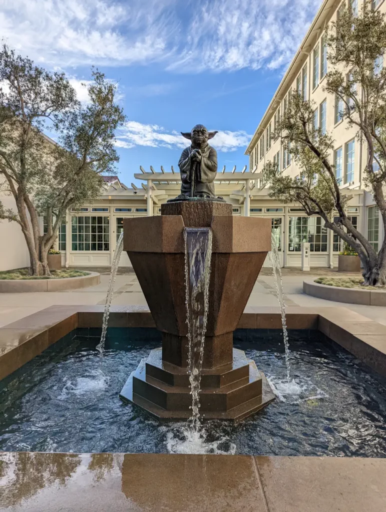 Fontaine vue de face, sur laquelle se trouve une statue de Maitre Yoda. Derrière se trouvent les locaux du siège social de Lucas Film.