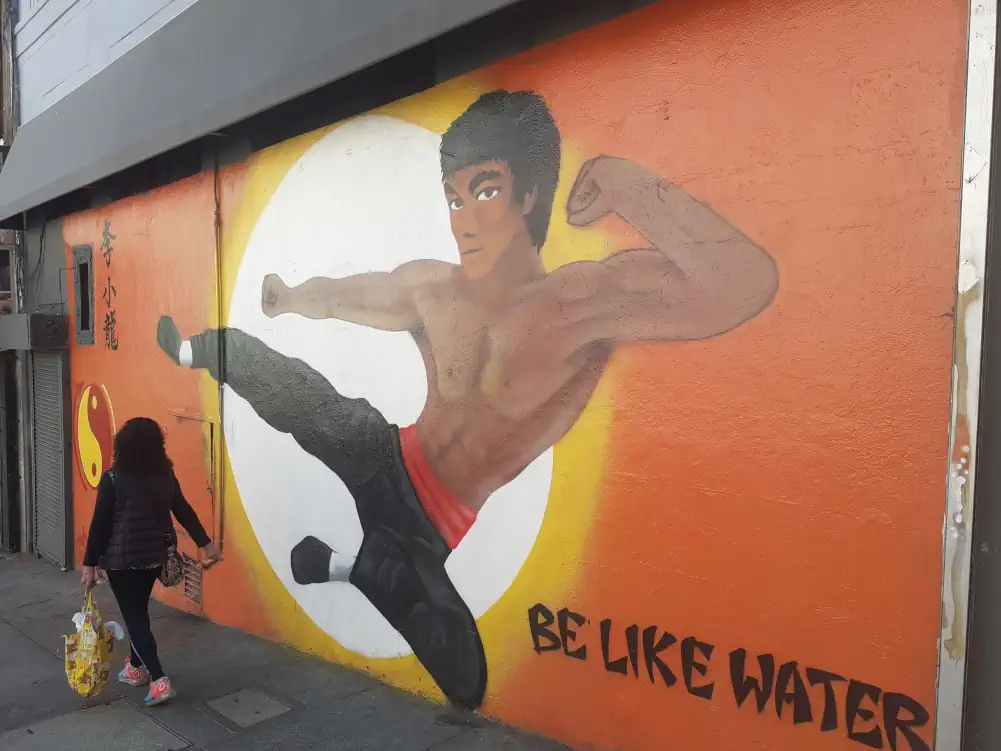 Fresque murale d'un Bruce Lee au corps disproportionné avec des petites jambes, devant un soleil et un fond orange.