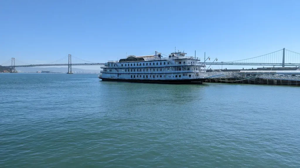 Navire blanc “San Francisco Belle”, flottant sur l’eau. Derrière se trouve un pont de San Francisco, le Oakland Bay Bridge.