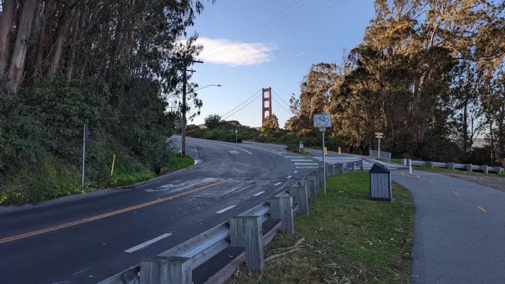 Au fond d’une route bordée par les arbres émerge le sommet d’un pylône rouge du pont de San Francisco, en plein virage.