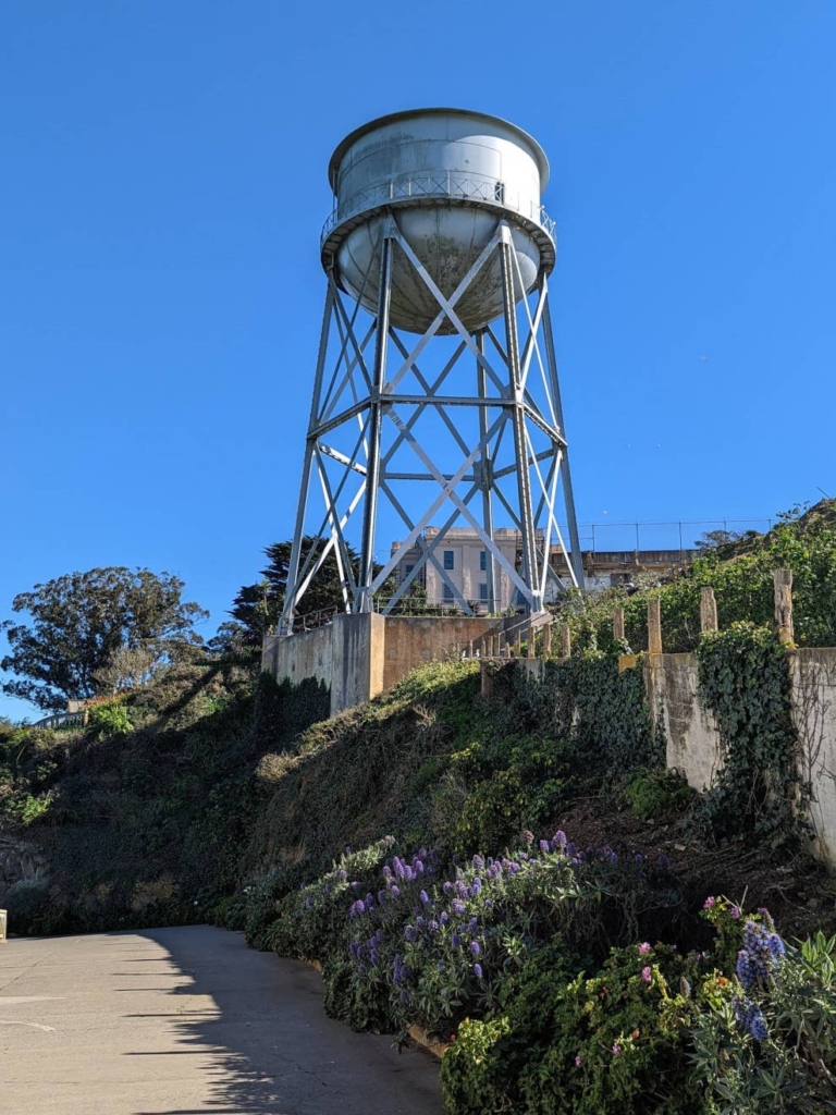 Contre-plongée sur le château d’eau d’Alcatraz. A ses pieds se trouve de la végétation verte et violette. Derrière se trouve un beau ciel bleu.