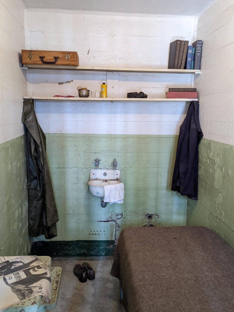 Intérieur d’une cellule de prisonnier. Elle est équipée d’un lit, d’un lavabo et de deux étagères. On y trouve également une valise en bois, quelques livres et une paire de chaussures.