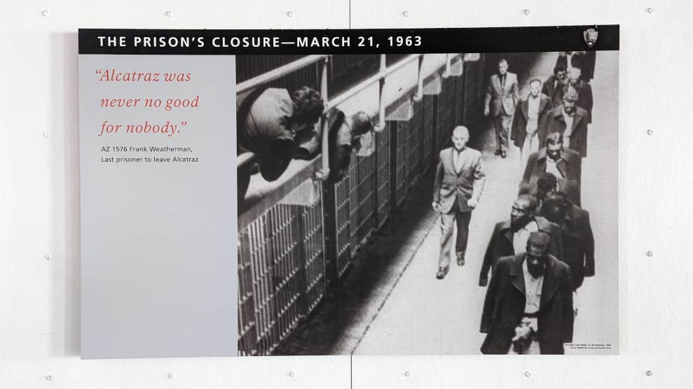 Photographie en noir et blanc de prisonniers longeant des cellules dans la prison d’Alcatraz. A gauche est inscrit le verbatim “Alcatraz was never good for nobody”.