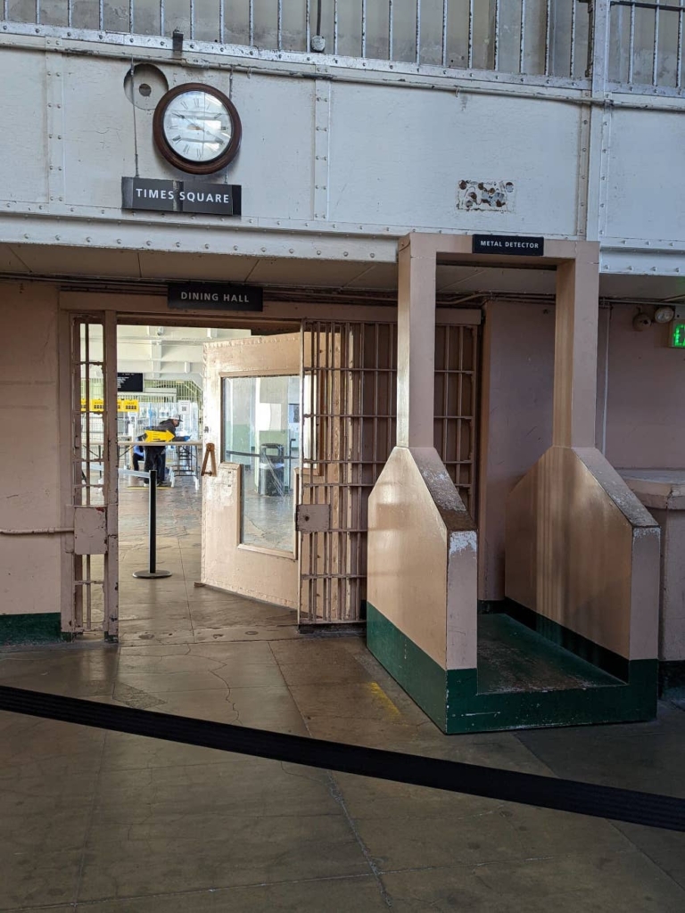 Entrée menant vers le réfectoire des prisonniers d’Alcatraz. A droite se trouve un détecteur de métal. Au-dessus se trouve une horloge et l’inscription “Times Square”.