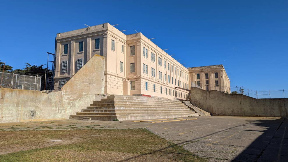 La prison d’Alcatraz, observée depuis la cour des prisonniers. Le bâtiment est accessible par des escaliers et éclairé par le soleil. En arrière-plan se trouve un ciel bleu sans nuages.