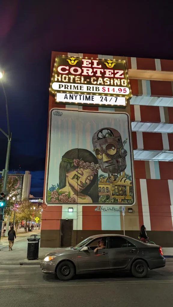 Peinture murale d’une femme portant une couronne à fleurs et d’un robot. Elle est située sous l’enseigne lumineuse de l’hôtel casino “El Cortez”.