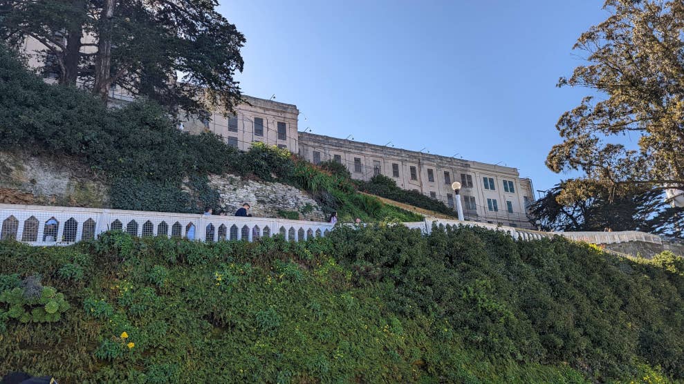 Vue en contre plongée sur la prison d’Alcatraz, devant un ciel bleu. Pour y accéder, nous pouvons emprunter un passage en pente entouré de végétation.