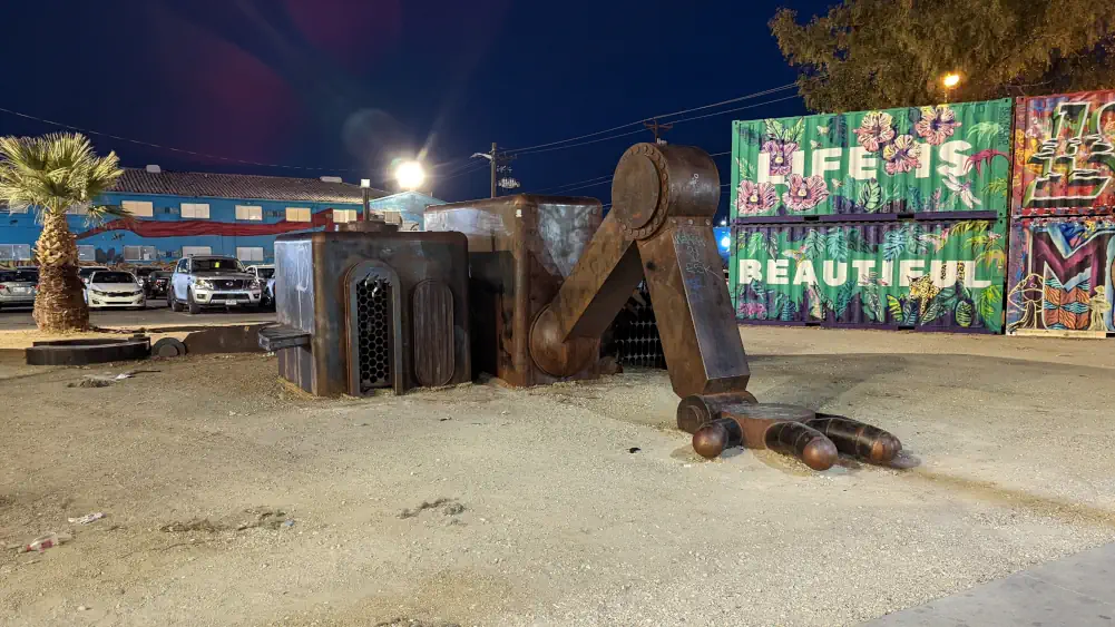 Sculpture d’un robot géant en bronze, face contre le sol sablé. Il semble vouloir se relever. Derrière lui se trouvent des containers peints.