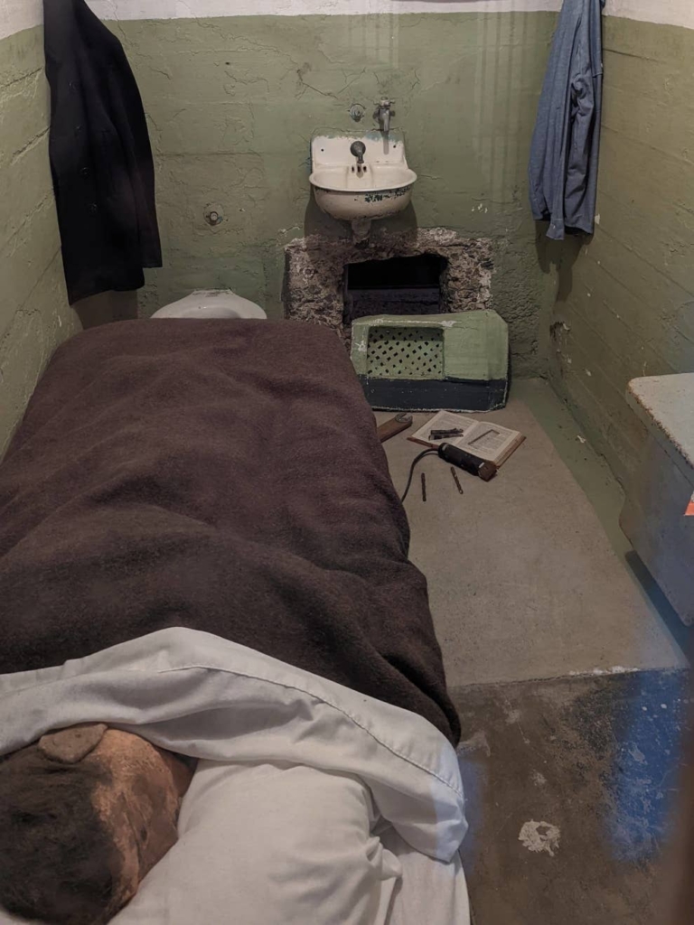 Intérieur d’une cellule. On observe un corps et une tête de mannequin au premier plan dans le lit. On observe également un trou sous le lavabo.