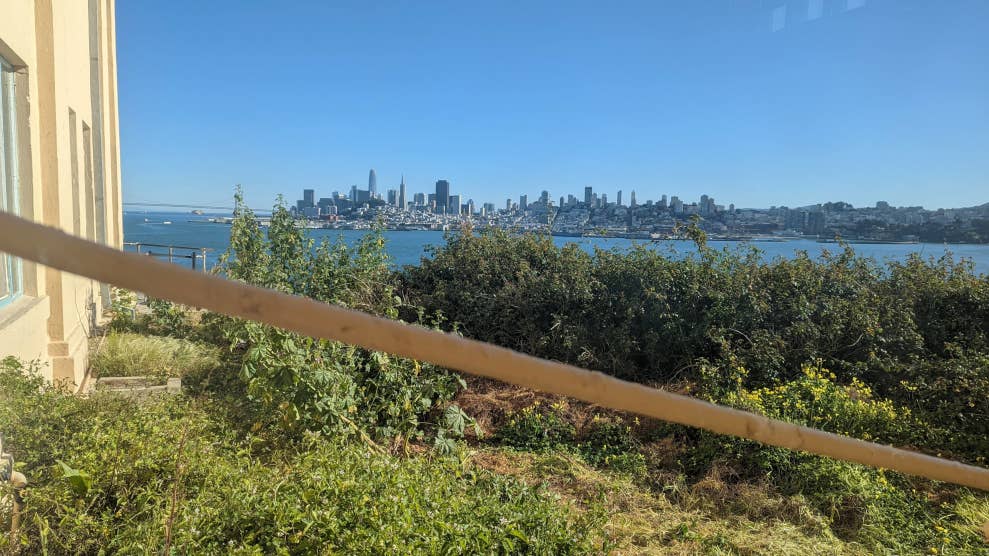 Vue d’un détenu observant à travers un hublot de la prison d’Alcatraz. On y voit de la végétation en premier et des gratte-ciel en arrière-plan.