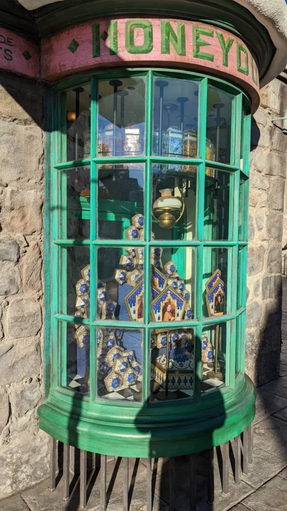 Vitrine aux rebords verts de la boutique “Honeydukes”. A travers les vitres, on aperçoit des boîtes de chocogrenouilles bien rangées.