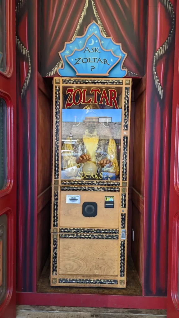 Fortune telling machine en bois Zoltar Speaks, plein pied. Sur le dessus se trouve un panneau bleu “Ask Zoltar?”. Le derrière est décoré d’un faux rideau rouge.