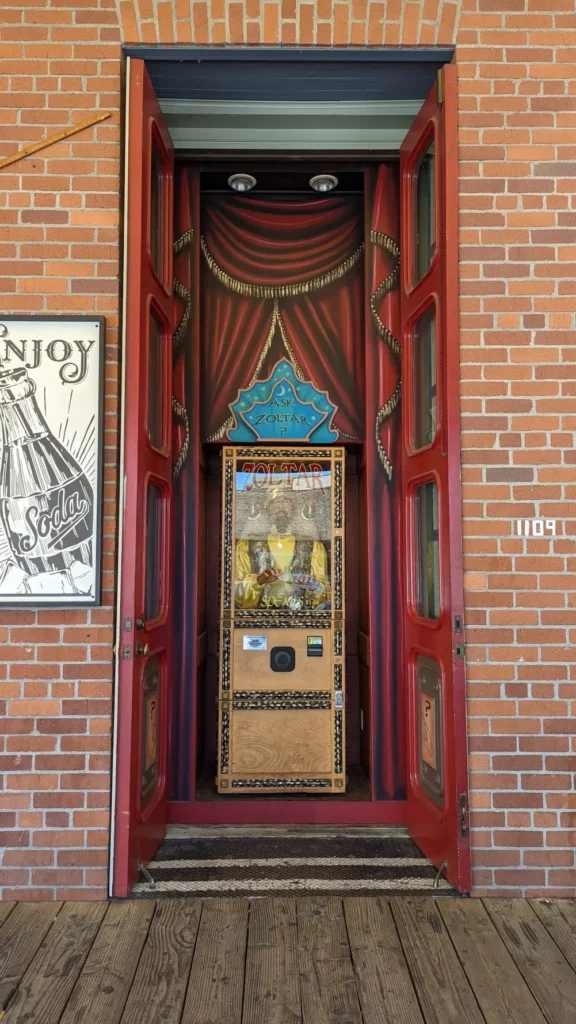 Face à face avec le renfoncement où se cache la fortune teller machine Zoltar Speaks. Sur les côtés se trouvent deux grandes portes rouges.