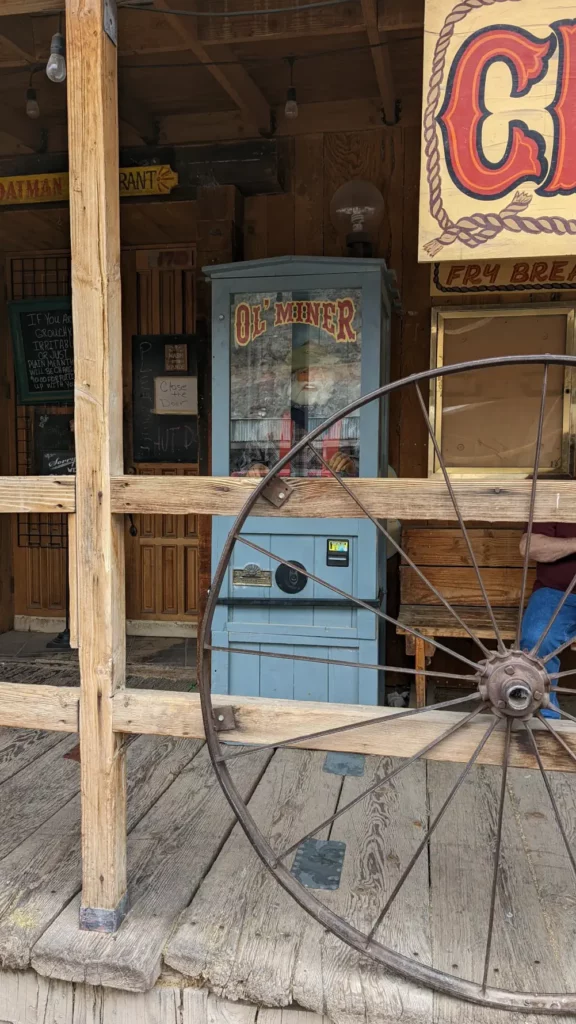 Fortune telling machine “Ol’Miner” bleue. Elle se cache derrière une rambarde en bois et une roue de calèche rouillée.