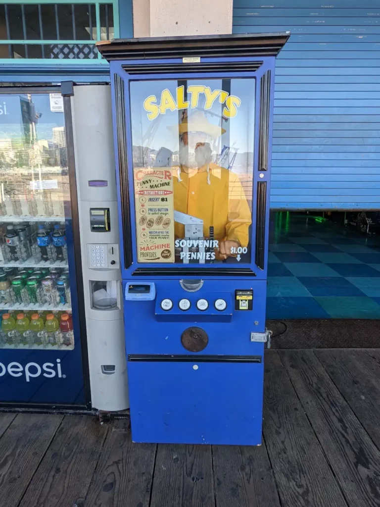 Portrait plein pied de la fortune telling machine bleu nuit “Salty’s”. A l’intérieur se trouve l’automate d’un vieil homme portant un ciré jaune.