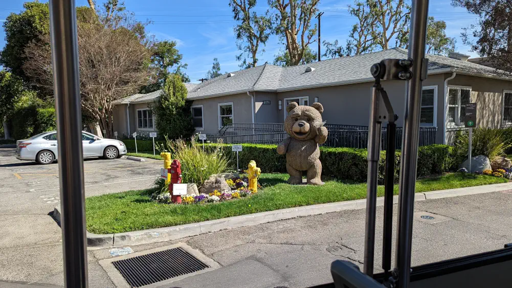 L’ours en peluche Ted se tenant debout dans l’herbe, devant une maison. Il salue les passagers du train en portant un toast.